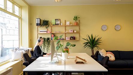 Bildet viser et kontorlokale med gule, kule vegger. Stor hvit kontorpult hvor det sitter en dame som jobber på laptop.